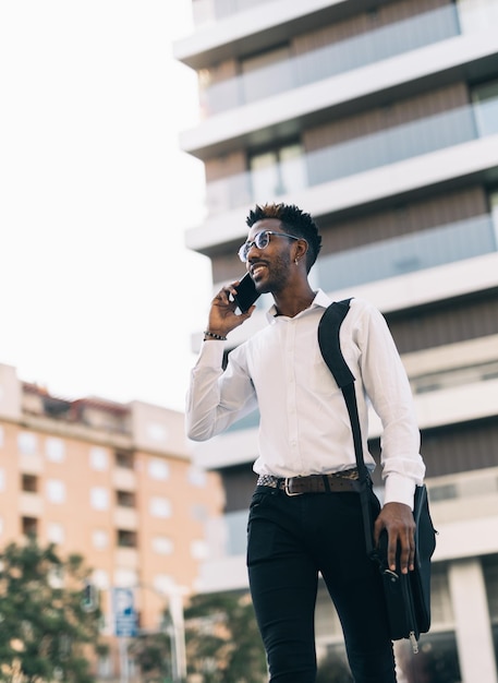 Souriant jeune homme d'affaires afro-américain marchant dans la rue en parlant sur son téléphone intelligent
