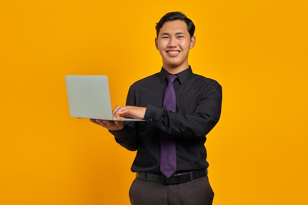 Souriant jeune homme d'affaires asiatique tenant et utilisant un ordinateur portable isolé sur fond jaune