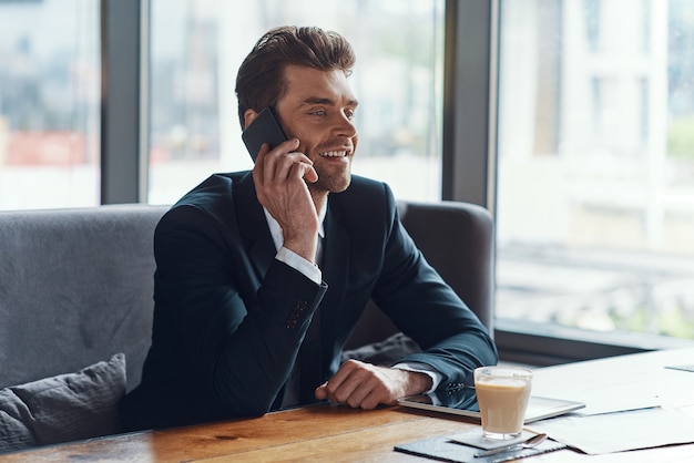 Souriant jeune homme d'affaires en costume complet parlant au téléphone intelligent alors qu'il était assis dans le restaurant