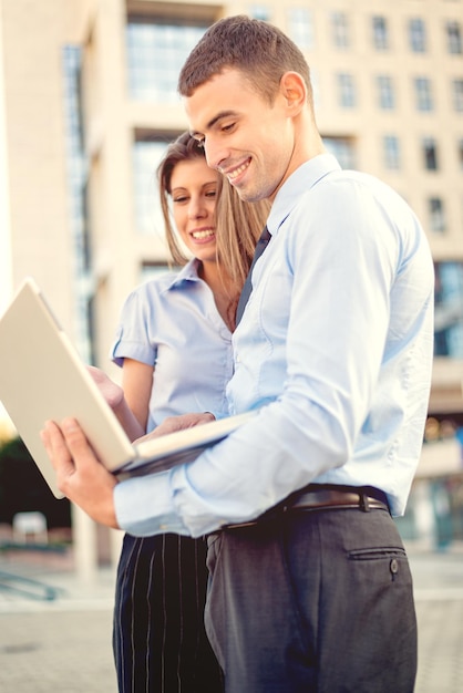 Souriant jeune homme d'affaires et femme d'affaires devant l'immeuble de bureaux regardant l'ordinateur portable, tandis que derrière le soleil brille.