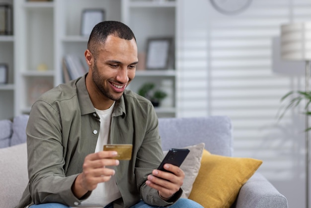 Souriant jeune homme afro-américain assis sur un canapé à la maison et utilisant une carte de crédit et une vérification par téléphone