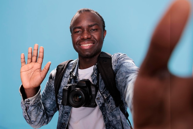 Souriant jeune homme agitant la caméra tout en prenant une photo de selfie sur fond bleu. Heureux photographe avec appareil DSLR et sac à dos de voyage prenant une photo de lui-même. Prise de vue en studio