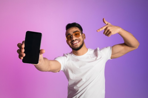 Souriant jeune homme arabe à lunettes de soleil pointant vers un smartphone avec un écran vide à la lumière du néon