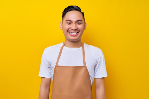 Souriant jeune homme asiatique des années 20 barista barman employé portant un tablier marron travaillant dans un café l'air confiant devant la caméra isolé sur fond jaune Concept de démarrage de petite entreprise