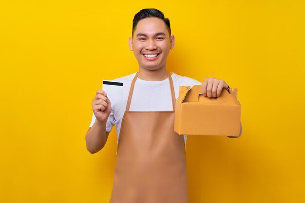 Souriant jeune homme asiatique barista barman employé portant un tablier marron travail dans un café tenant un sac à gâteau en papier à emporter et une carte bancaire de crédit sur fond jaune Concept de démarrage de petite entreprise