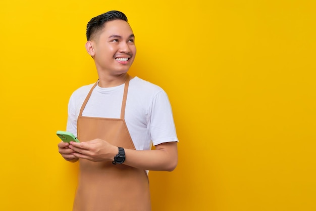 Souriant jeune homme asiatique barista barman employé portant un tablier marron travail dans un café tenant un téléphone portable et regardant de côté isolé sur fond jaune Concept de démarrage de petite entreprise