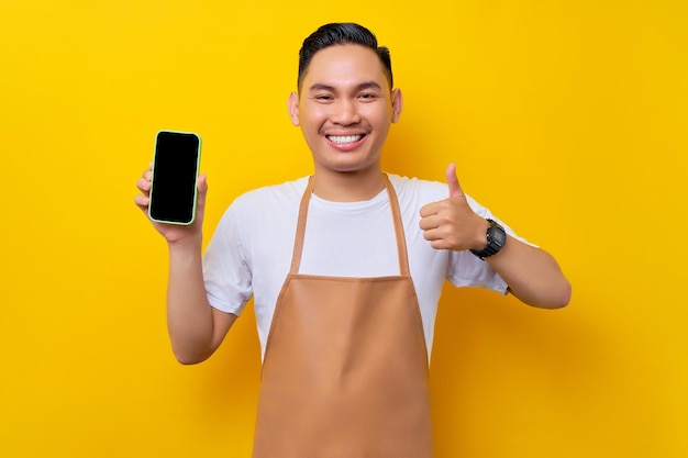 Souriant jeune homme asiatique barista barman employé portant un tablier marron travaillant dans un café montrant une maquette d'écran vide de téléphone portable et un signe de pouce vers le haut sur fond jaune Démarrage d'une petite entreprise