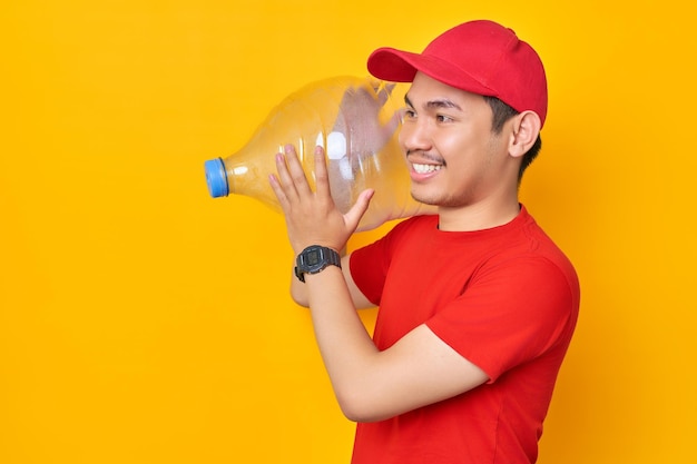 Souriant jeune homme asiatique en bonnet rouge tshirt uniforme employé travailler comme concessionnaire courrier tenant une bouteille d'eau à la recherche de côté isolé sur fond jaune Concept de service de livraison professionnel