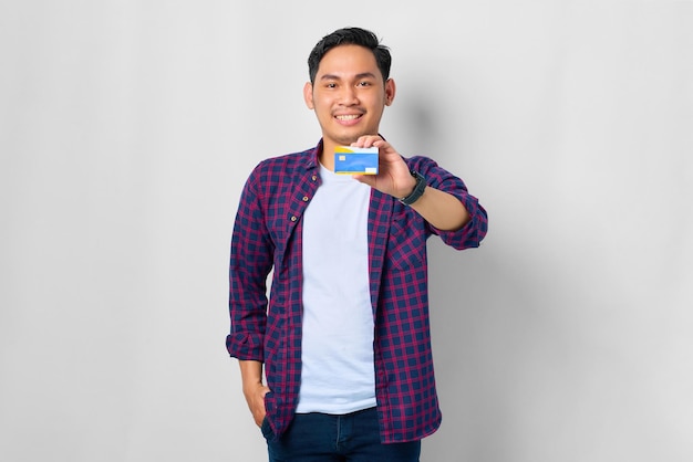 Souriant jeune homme asiatique en chemise à carreaux montrant la carte de crédit isolé sur fond blanc