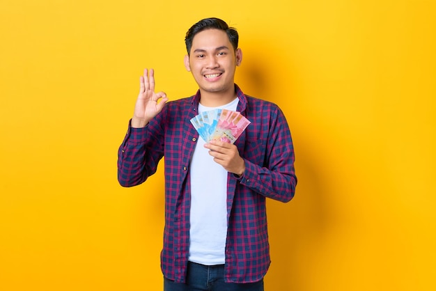 Souriant jeune homme asiatique en chemise à carreaux tenant des billets de banque et gesticulant signe d'accord isolé sur fond jaune