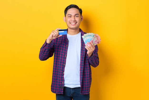 Souriant jeune homme asiatique en chemise à carreaux tenant des billets de banque et montrant une carte de crédit isolée sur fond jaune