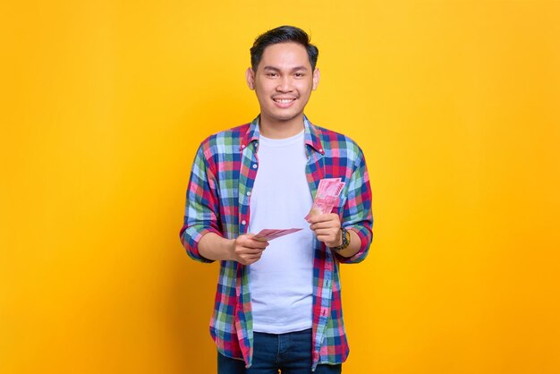 Souriant jeune homme asiatique en chemise à carreaux tenant des billets de banque et regardant la caméra isolée sur fond jaune