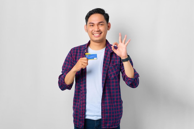 Souriant jeune homme asiatique en chemise à carreaux tenant une carte de crédit et gesticulant signe d'accord isolé sur fond blanc