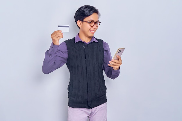 Souriant jeune homme asiatique en chemise décontractée et gilet à l'aide de smartphone et carte de crédit isolé sur fond blanc Concept de style de vie des gens