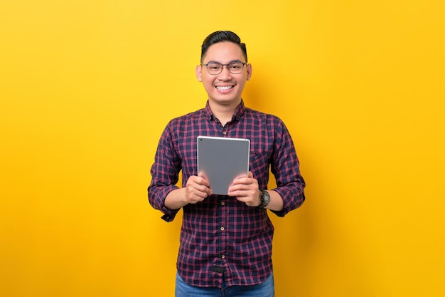 Souriant jeune homme asiatique dans des verres tenant une tablette numérique et regardant la caméra isolée sur fond jaune