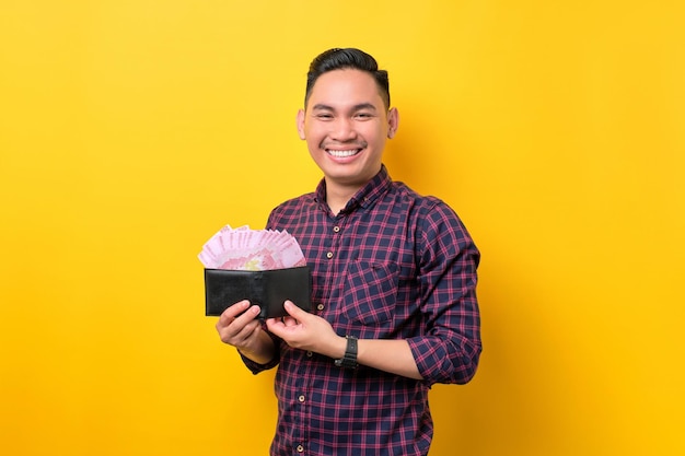 Souriant jeune homme asiatique montrant un portefeuille plein de billets de banque isolés sur fond jaune Concept de profit et de richesse