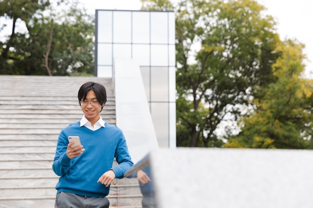 Souriant jeune homme asiatique parlant au téléphone mobile en se tenant debout à l'extérieur de l'escalier