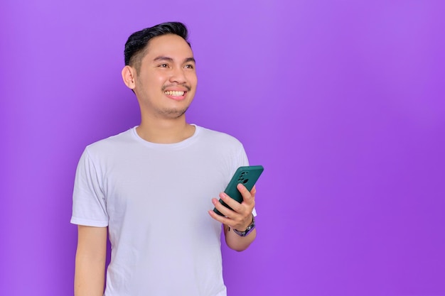 Souriant jeune homme asiatique en t-shirt blanc utilisant un smartphone et regardant loin l'espace de copie isolé sur fond violet