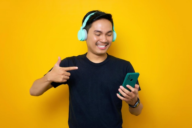 Souriant jeune homme asiatique en t-shirt décontracté avec un casque sans fil pointant du doigt un téléphone portable isolé sur fond jaune Concept de mode de vie des émotions des gens