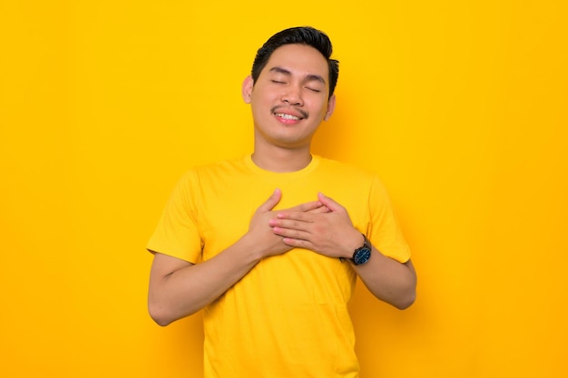 Souriant jeune homme asiatique en t-shirt décontracté tenant les mains sur sa poitrine isolé sur fond jaune Concept de style de vie des gens