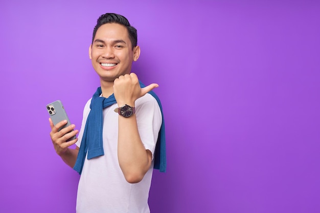 Souriant jeune homme asiatique en t-shirt décontracté tenant un téléphone portable et pointant le pouce de côté sur l'espace de copie isolé sur fond violet