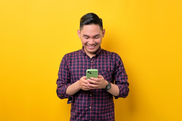 Souriant jeune homme asiatique utilisant un smartphone pour envoyer des messages avec des amis isolés sur fond jaune