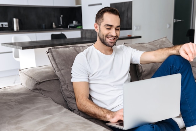 Souriant jeune homme assis sur un canapé à la maison, à l'aide d'un ordinateur portable