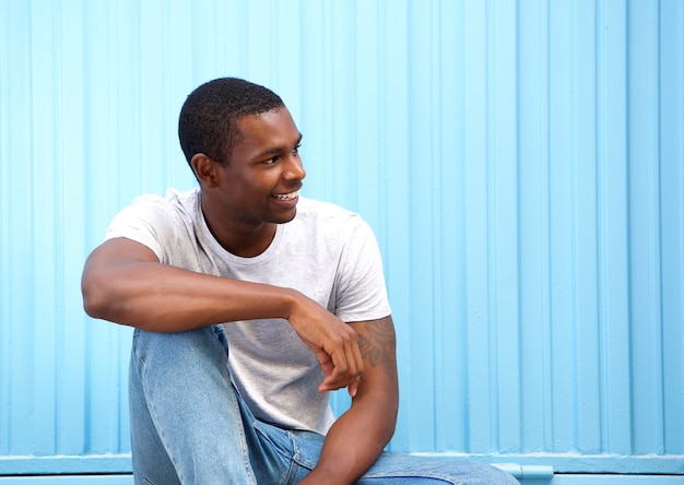 Photo souriant jeune homme assis contre le mur bleu en détournant les yeux