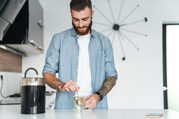 Souriant jeune homme barbu faisant une tasse de thé en se tenant debout dans la cuisine à la maison