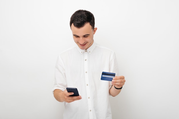 Souriant jeune homme en blanc sur blanc en regardant le téléphone et tenant sa carte de crédit. Services bancaires mobiles et concept de magasinage en ligne.