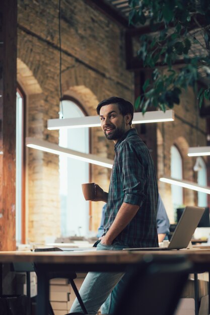 Souriant jeune homme en chemise à carreaux debout dans le bureau avec son dos à la table et tenant une tasse