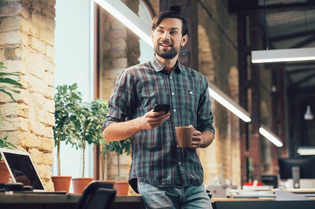 Souriant jeune homme debout près de la table avec une tasse et un smartphone et détournant les yeux