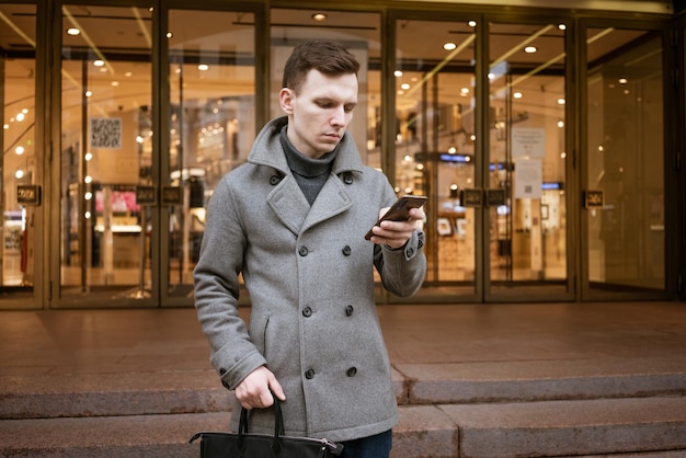 Souriant jeune homme en manteau gris tient des messages texte défilants dans son mobile