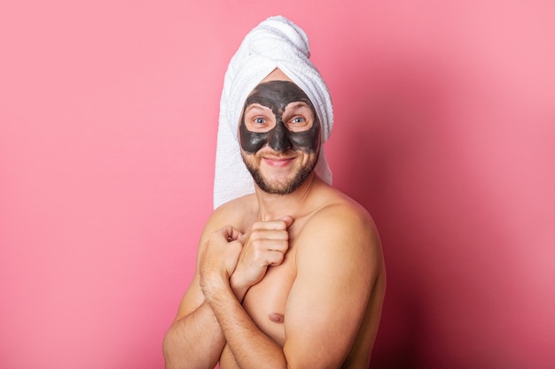 Souriant jeune homme avec masque cosmétique, nu se cachant derrière sur fond rose.