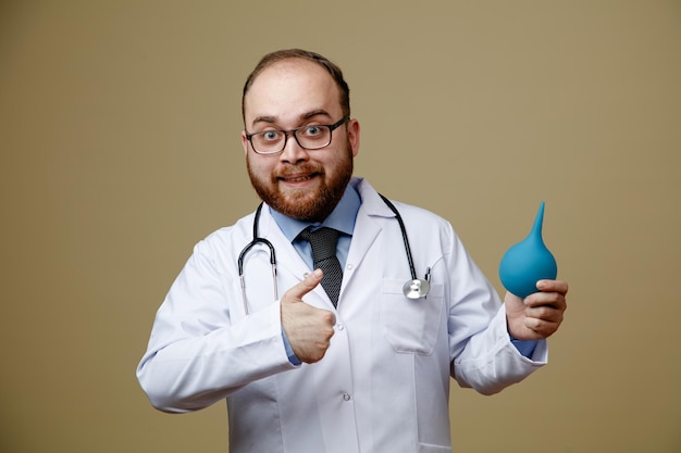 Souriant jeune homme médecin portant des lunettes blouse de laboratoire et stéthoscope autour du cou tenant un lavement regardant la caméra montrant le pouce vers le haut isolé sur fond vert olive