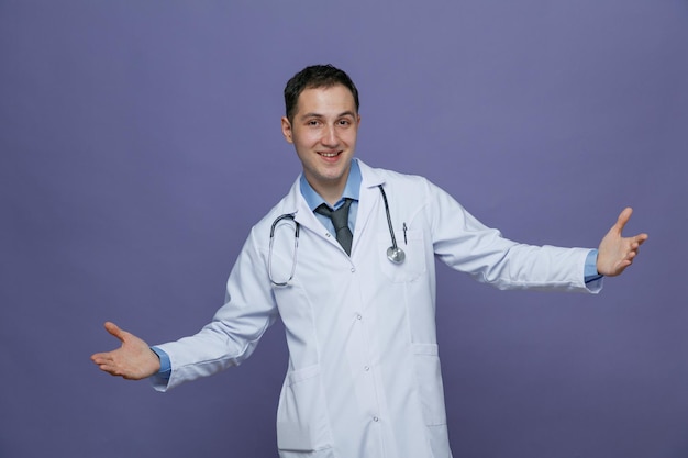 Souriant jeune homme médecin portant une robe médicale et un stéthoscope autour du cou regardant la caméra montrant les mains vides avec les bras grands ouverts isolés sur fond violet