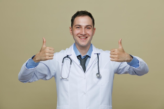 Souriant jeune homme médecin portant une robe médicale et un stéthoscope autour du cou regardant la caméra montrant les pouces vers le haut isolé sur fond vert olive