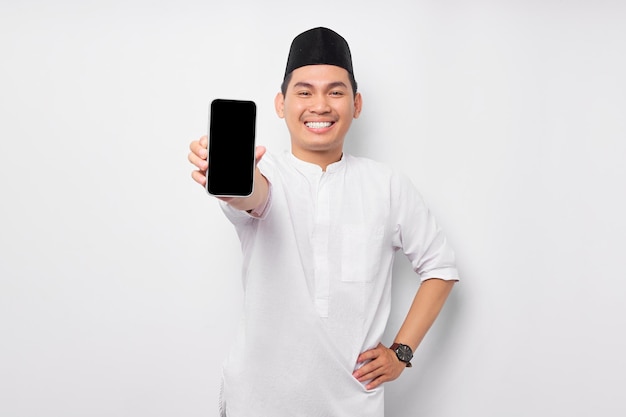 Souriant jeune homme musulman asiatique montrant l'écran du téléphone portable et la main sur sa taille isolé sur fond blanc Les gens religieux Islam style de vie concept célébration Ramadan et ied Moubarak