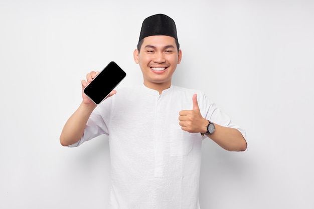 Souriant jeune homme musulman asiatique montrant l'écran du téléphone portable montrant le geste du pouce vers le haut isolé sur fond blanc Les gens religieux Islam style de vie concept célébration Ramadan et ied Moubarak