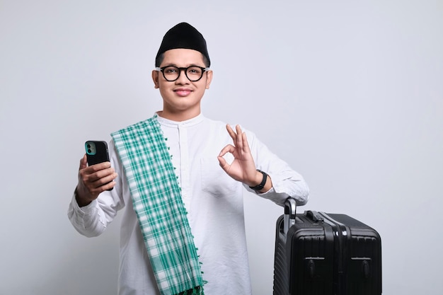 Souriant jeune homme musulman asiatique montrant un signe ok tout en tenant un smartphone et en s'appuyant sur une valise noire