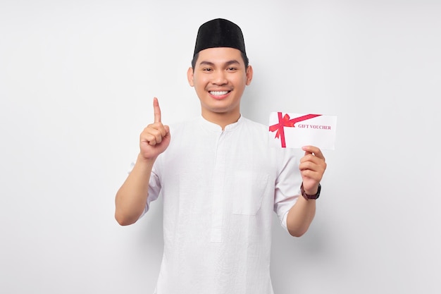 Souriant jeune homme musulman asiatique tenant un bon de certificat-cadeau pointant son doigt vers le haut isolé sur fond blanc Les gens religieux Islam style de vie concept célébration Ramadan et ied Moubarak