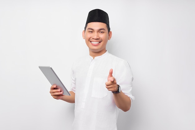 Souriant jeune homme musulman asiatique tenant une tablette numérique et pointant le doigt sur la caméra isolé sur fond blanc Les gens religieux Islam style de vie concept célébration Ramadan et ied Moubarak
