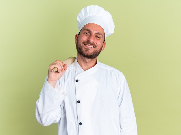 Souriant jeune homme de race blanche cuisinier en uniforme de chef et casquette tenant un rouleau à pâtisserie sur l'épaule