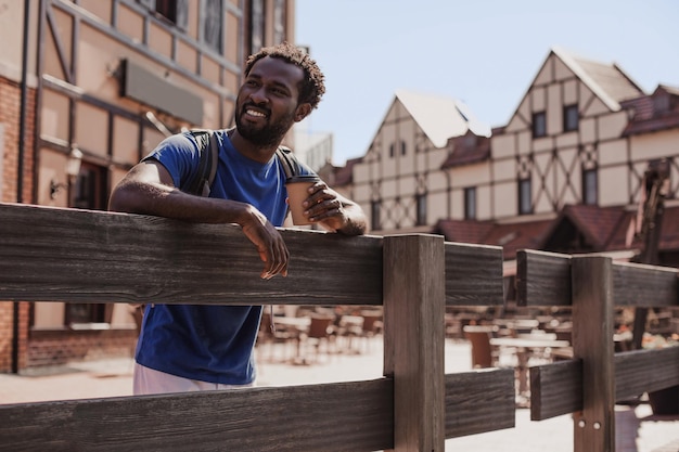 Souriant jeune homme avec sac à dos et tasse de café debout à une clôture en bois