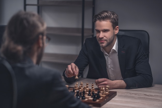 Souriant jeune homme séduisant intelligent avec pièce d'échecs en main et adversaire jouant au bureau