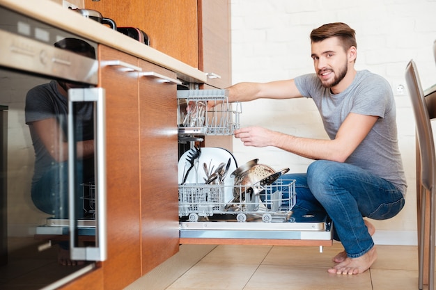 Souriant jeune homme séduisant utilisant un lave-vaisselle dans la cuisine à la maison