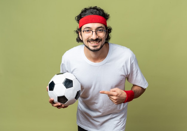 Souriant jeune homme sportif portant un bandeau avec un bracelet tenant et pointe au ballon