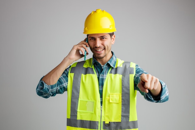 Souriant jeune ingénieur masculin portant un casque de sécurité et un uniforme regardant et pointant vers la caméra tout en parlant au téléphone isolé sur fond blanc