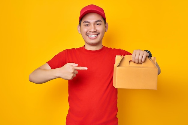 Souriant jeune livreur asiatique en vêtements de travail de t-shirt à casquette rouge travaille comme courrier de revendeur pointant du doigt le dessert de gâteau dans une boîte vide non marquée isolée sur fond jaune Concept de service