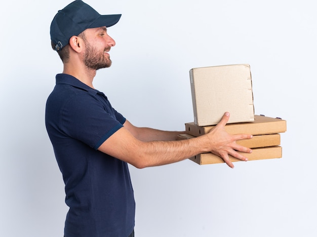 Souriant jeune livreur caucasien en uniforme bleu et casquette debout en vue de profil étirant les paquets de pizza et la boîte en carton les regardant